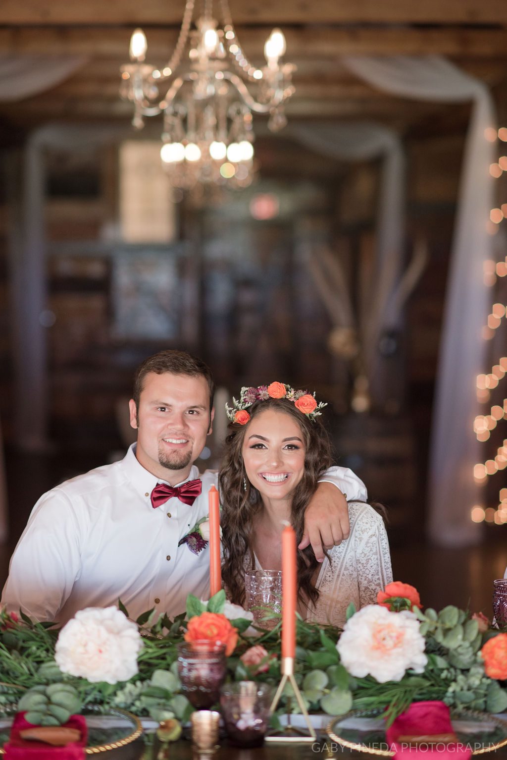 Bride wearing floral crown and groom wearing crimson bowtie sitting at sweetheart table