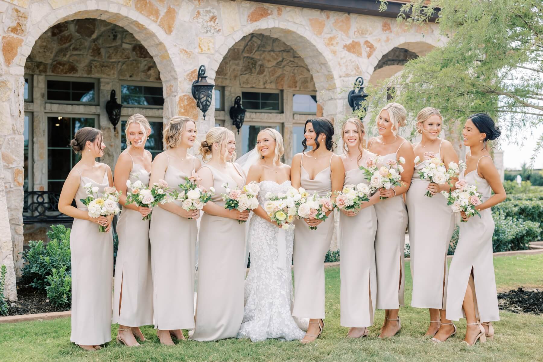 Bride standing with bridesmaids in champagne dresses with white and pastel bouquets 