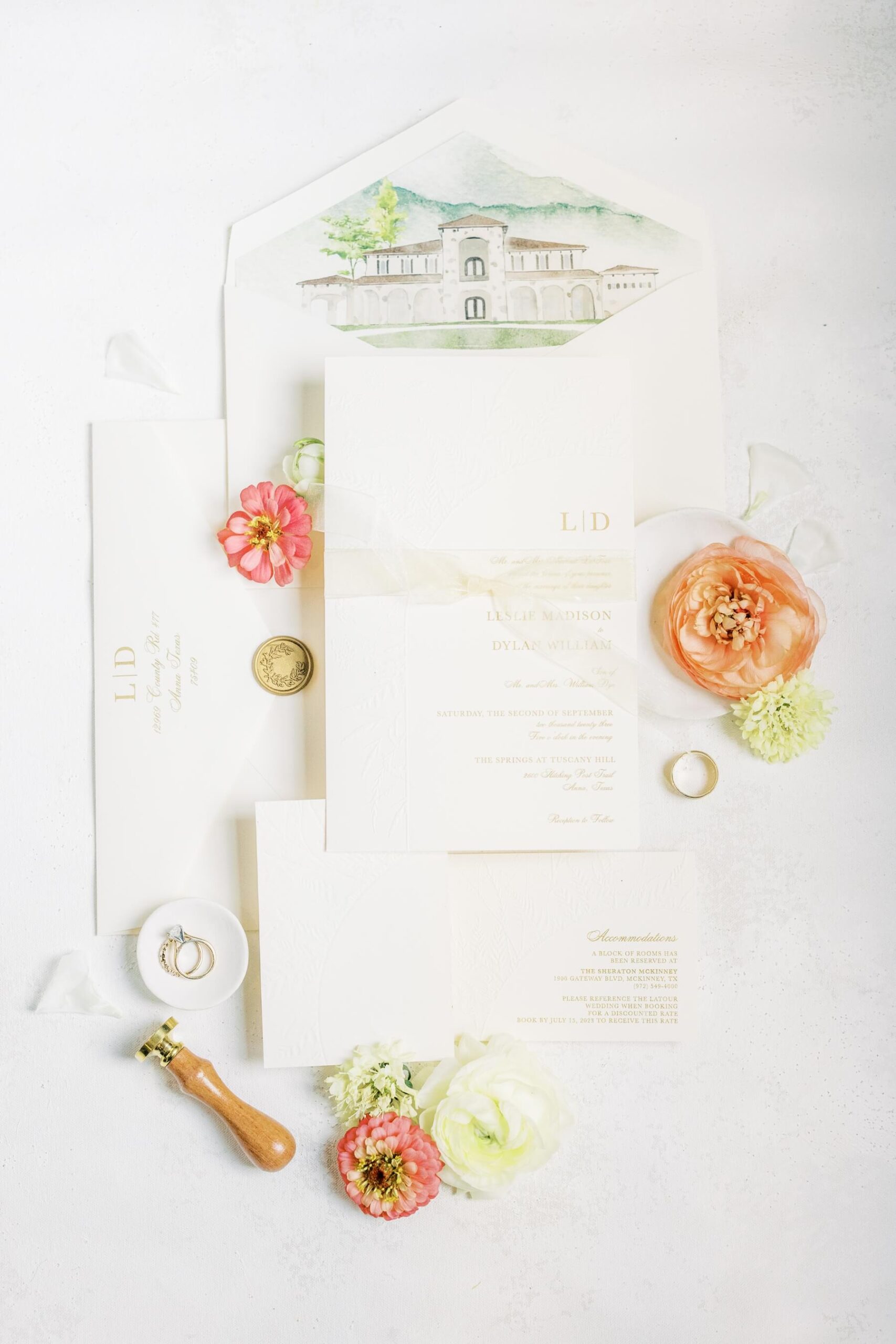 Wedding invitation flat lay featuring white invitations with gold font, wedding rings, and pink and white flowers