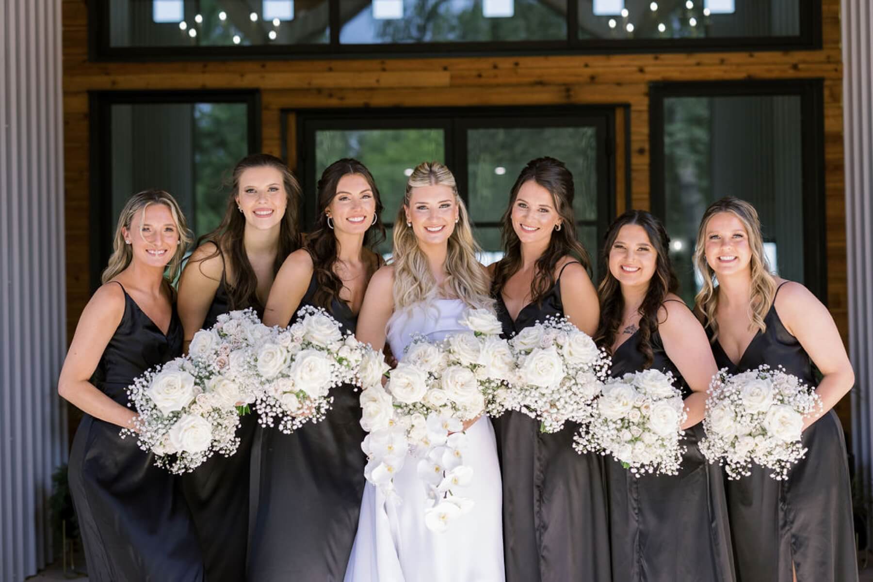 Bride holding bouquet with white orchids, roses, and baby's breath with bridesmaids in black dresses holding white bouquets at Union House TX