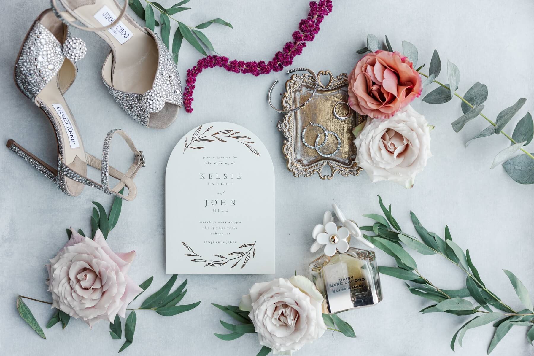 Flat lay featuring white wedding invitation, silver bridal shoes, perfume, and bride's jewelry