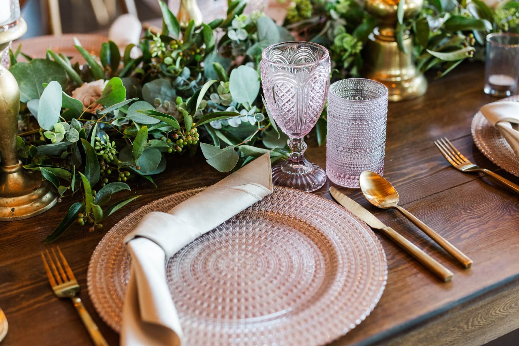 Pink charger with pink glassware, gold place settings, and greenery runners