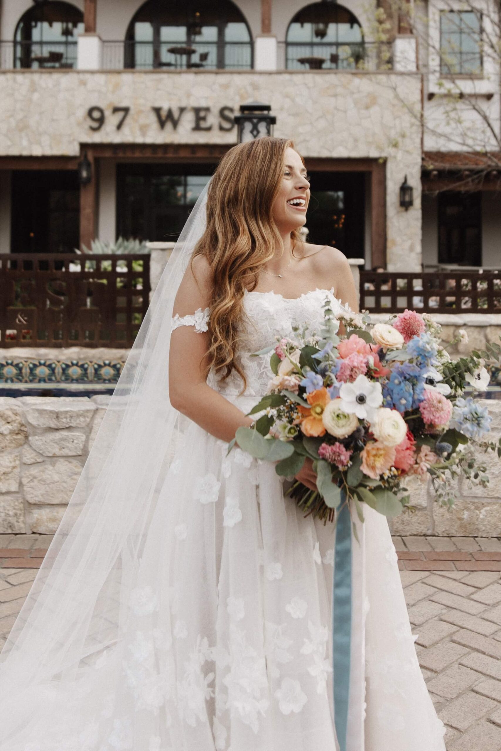 Bride holding bouquet with pink, blue, orange, and white flowers in front of Hotel Drover