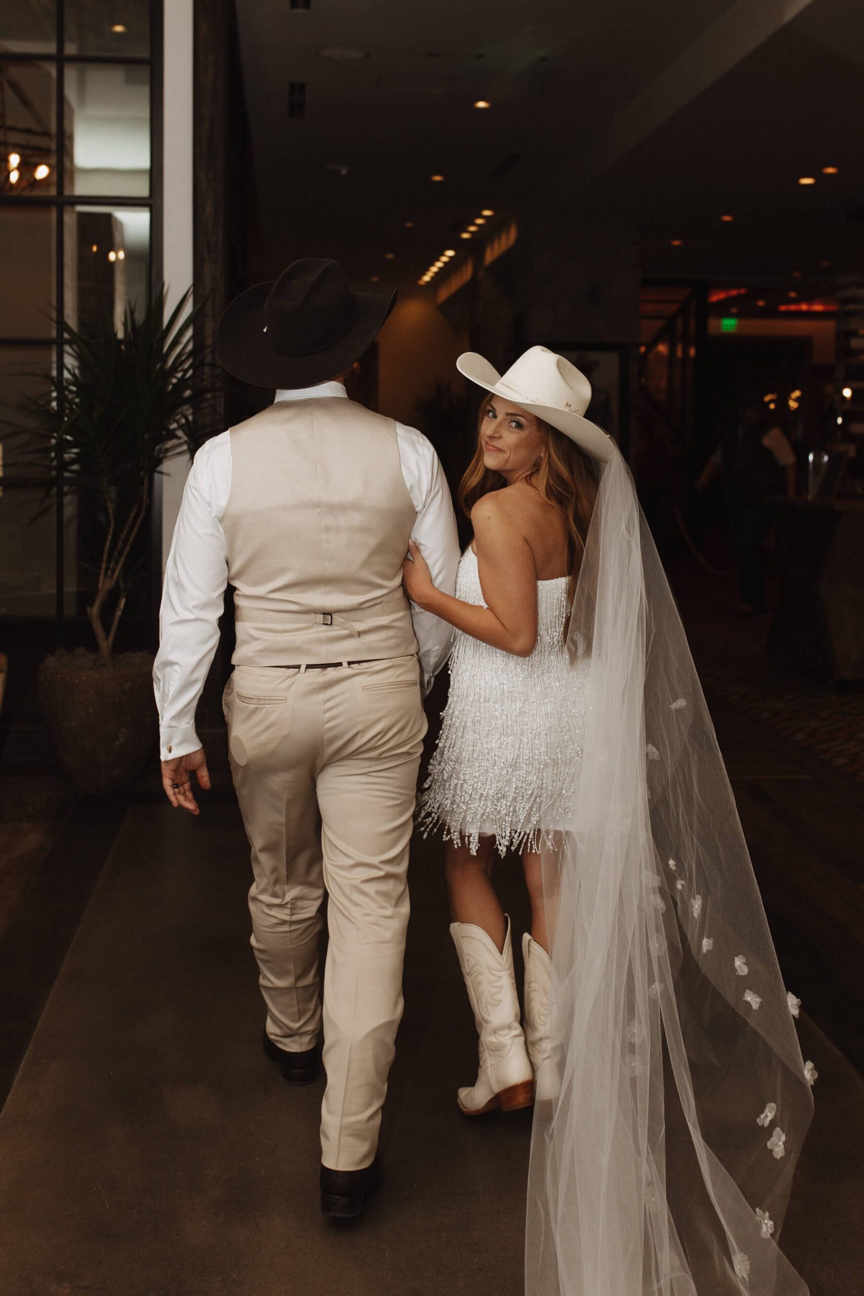 Bride wearing white cowgirl hat with veil, short white dress, and white boots walking with groom